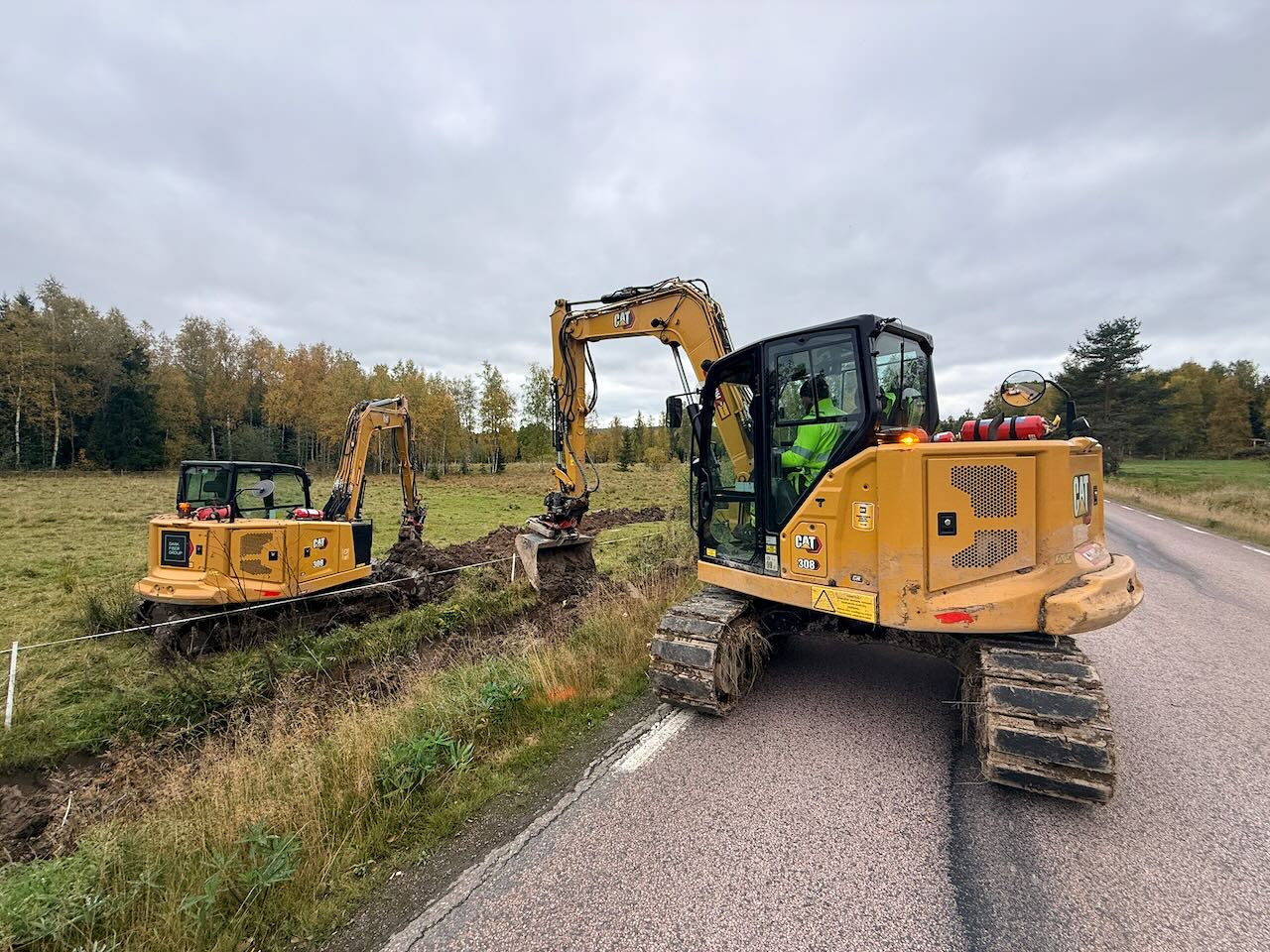 Two excavators digging a fiber duct trench alongside a rural Nordic road